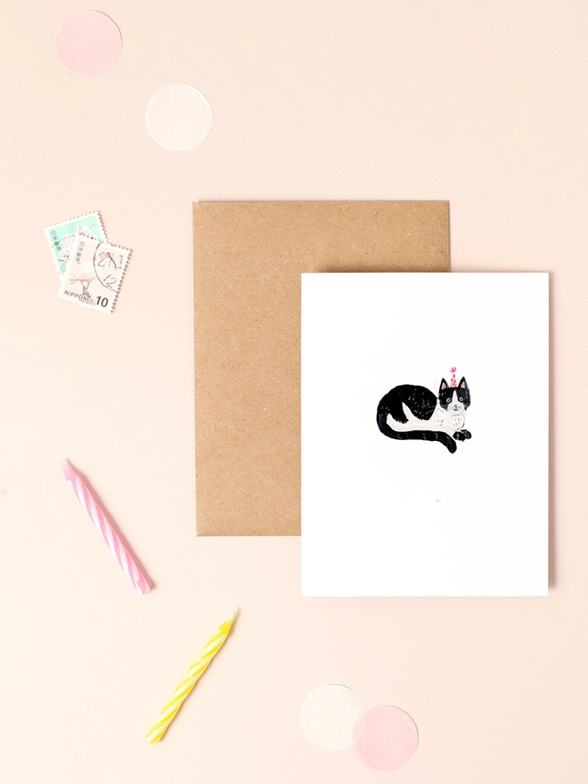 An image of a mini, rectangular white greetings card with an illustration of a black and white cat wearing a party hat. The mini greetings card is placed on top of a rectangular brown kraft envelope.