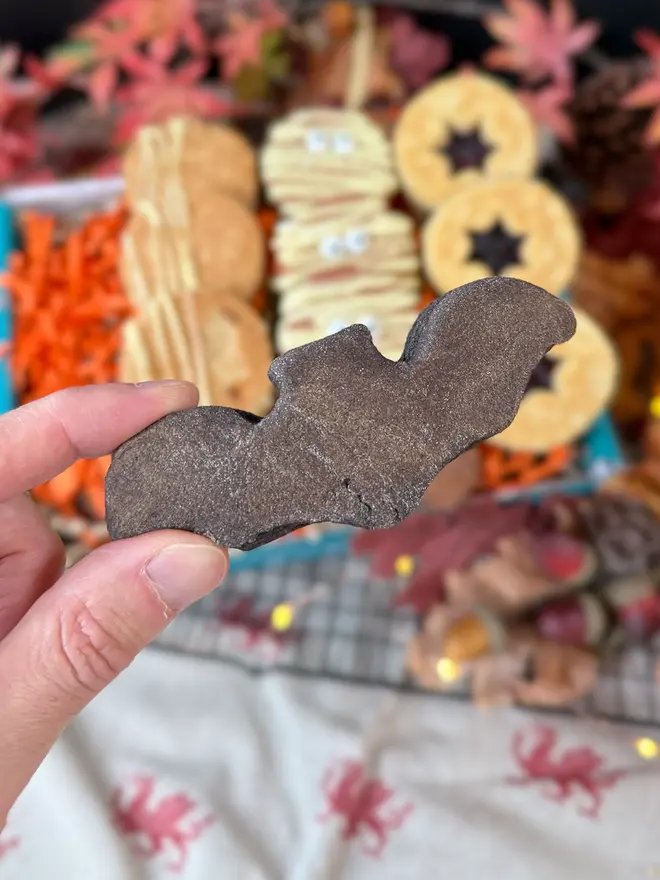 A hand holds a black velvet bat-shaped welshcake in the foreground, with a blurred background of Halloween-themed welshcakes and autumn decorations.
