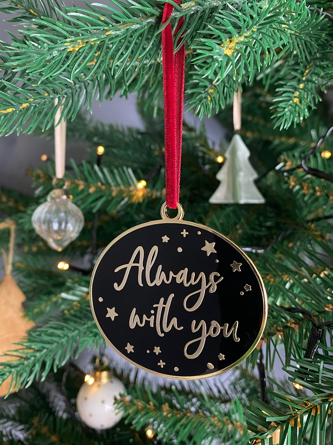 A black and gold enamel Christmas decoration, with the words “Always With You” surrounded by gold stars, hangs on a Christmas tree.