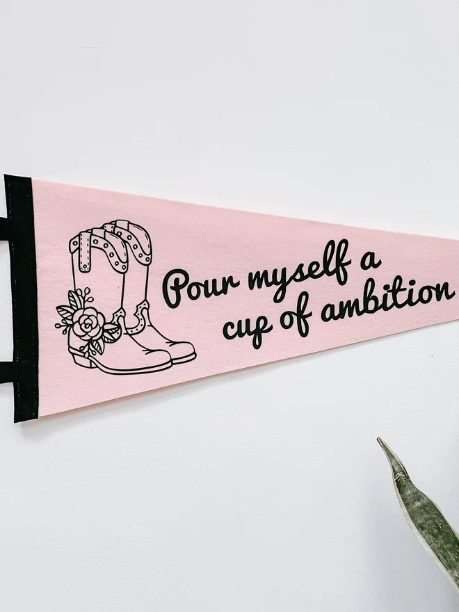 a pastel pink felt pennant flag with an image of a pair of cowboy boots and the phrase 'pour myself a cup of ambition' from Dolly Parton's song 9 to 5