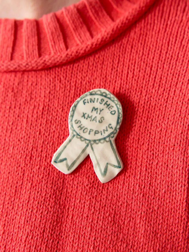 'Finished my Xmas Shopping' hand-painted on a ceramic Christmas award badge in green, pinned to a woolen red knitted jumper