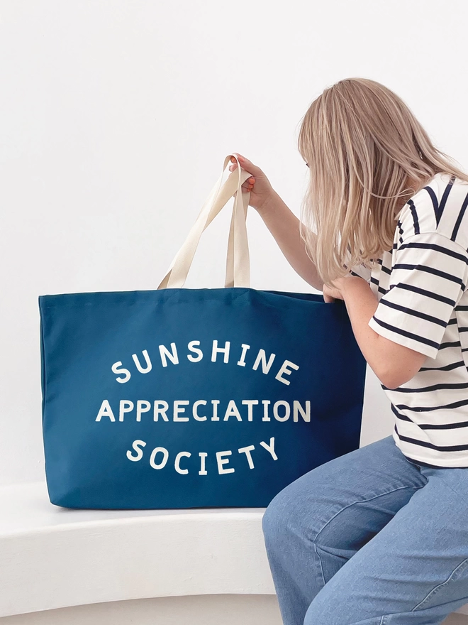 Blue oversized sunshine appreciation society bag sitting on a bench with model peering inside