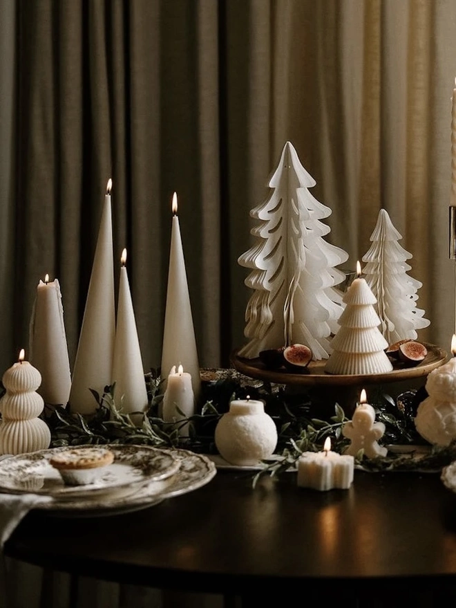 White Christmas Tree Cone Candles (Set Of 3). A festive dinner table with tall cream cone candles, ribbed tree and bauble candles, a gingerbread man candle and paper trees, styled with greenery and figs. The mix of sculptural candles creates a warm, sophisticated holiday tablescape.