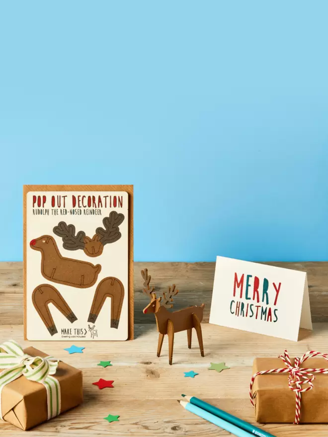 Pop out laser-cut Rudolph Christmas decoration and Merry Christmas card and brown kraft envelope on top of a wooden desk in front of a sky blue coloured background