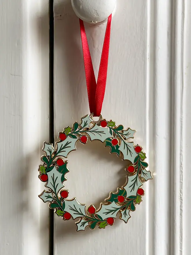 An heirloom Christmas wreath decoration with holly leaves and red berries hangs from a red ribbon on a white door handle.