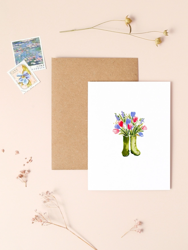 A charming illustration of a pair of green wellington boots filled with a vibrant bouquet of wild spring flowers, including pink tulips, bluebells, and yellow daffodils. The boots are set against a soft, neutral background, highlighting the colorful arrangement. The design exudes a rustic, countryside charm, perfect for conveying warmth and appreciation.