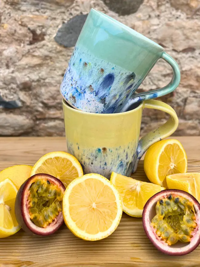 Shoreline Standard Mug, a pair of colourful mugs placed on a wooden surface stacked on top of each other. They are accompanied by fruit. 