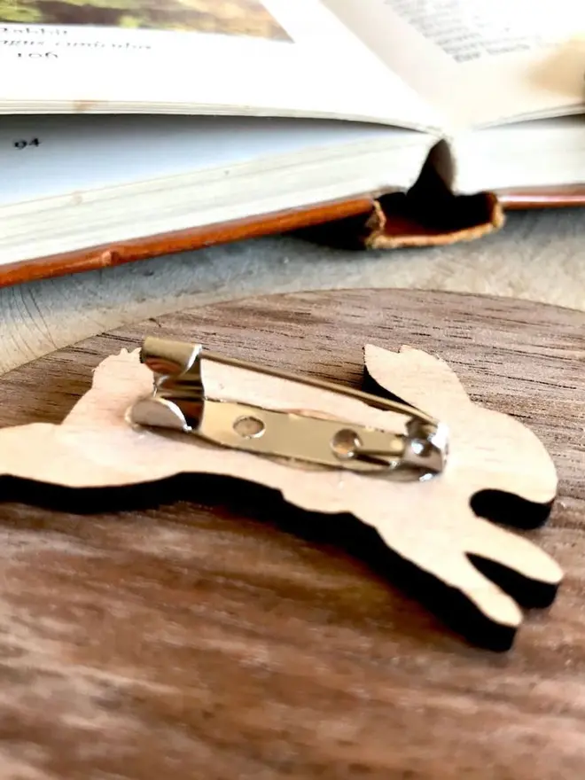 Birthday Girl Wooden Bear Badge, The back of a wooden rabbit brooch showing a metal safety pin fastener attached to the wooden cutout, resting on a round wooden surface in front of an open book.