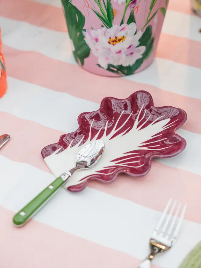 Radicchio Leaf Spoon Rest in Verona Red as part of a larger tablescape with a pink and white tablecloth, painted plant pots and colourful cutlery