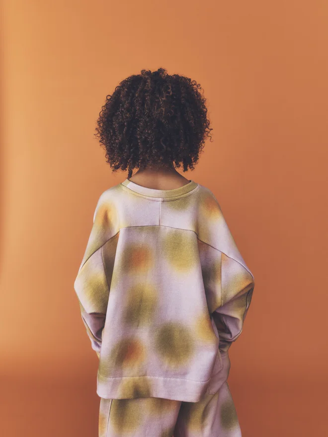 ack view of a child with hands behind their back, showing the yoke seam and dropped shoulders of the Studio Koter Riverson top in Hazy Lilac, worn with matching joggers.