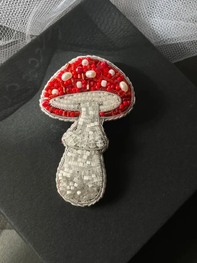 Mushroom Beaded Brooch displayed on a grey box, next to some white fabric. 