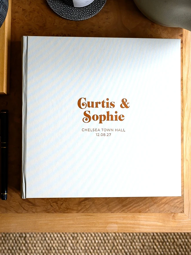 A pearl-coloured wedding guest book with gold printing on the cover rests on a wooden table.
