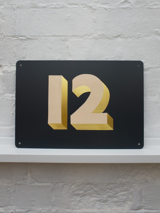 Hand painted peach and gold leaf house number 12 on an anthracite grey metal plaque against a white brick wall. 