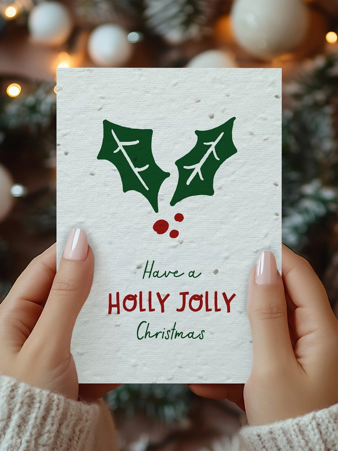A pair of hands holding a Christmas card with an illustrated holly leaf on it with 'Have a Holly Jolly Christmas' underneath
