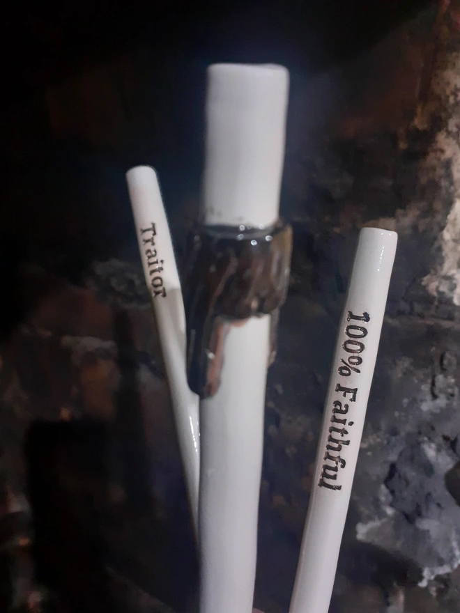 Handmade ceramic drinking straws inspired by The Traitors, featuring the words “Traitor” and “100% Faithful” alongside a sculptural dark fringe motif, glazed in white and black.