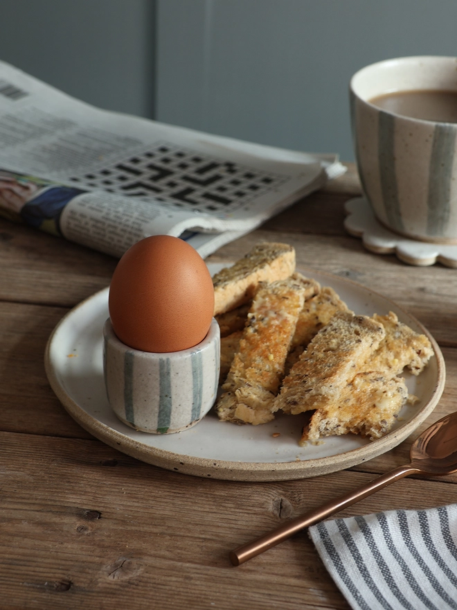 A breakfast scene featuring a handmade ceramic egg cups with a blue and white stripe design holding a boiled egg. The egg cup sits on a white handmade ceramic plate filled with buttered toast. A newspaper, cloth napkin, brass spoon and blue and white striped mug filled with coffee also sit on the table. 