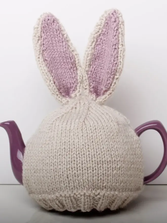 Hand Knitted Bunny Rabbit Tea Cosy, a rabbit bunny tea cosy sitting on a grey surface against a white backdrop. It fits over a purple tea pot. 