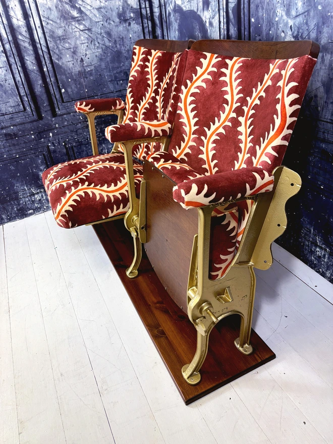 Side on view of two refurbished vintage cinema seats upholstered in 'Helter Skelter' redcurrant coloured velvet.  The flip down part of the seats are open.  The seats are mounted onto a wooden plinth in front of a blue marbled wall.