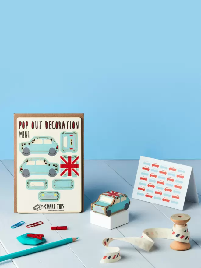 Pop out Mini car decoration and car pattern greeting card and brown kraft envelope on top of a wooden desk in front of a blue coloured background