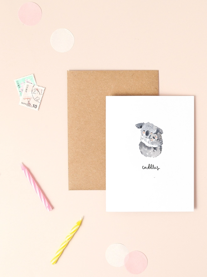 An image of a mini, rectangular white greetings card with an illustration of a couple of cuddling koalas with a hand written 'cuddles' message in black ink seen under the central illustration. The card is seen on top of a brown rectangular kraft envelope.