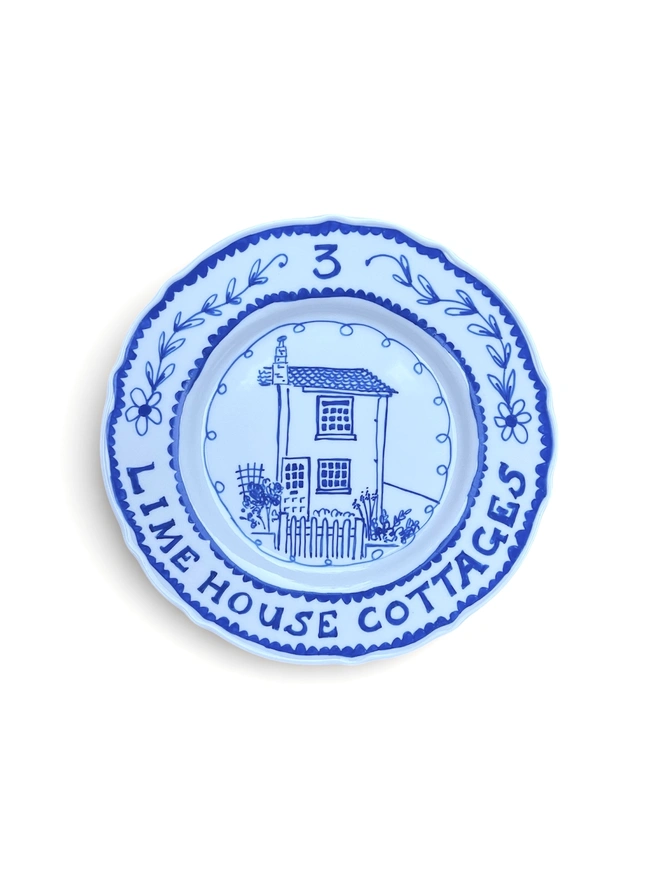 Blue and white plate, in a cartoon illustration style depicts an end terrace house with the address around the rim and a botanical illustration