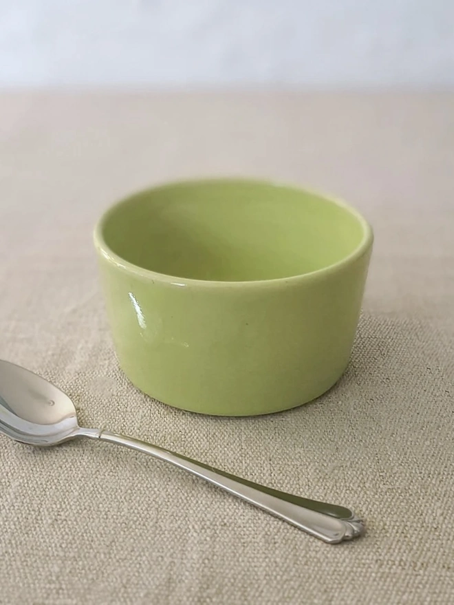 Classic Side Bowl, a colourful ceramic bowl on a wooden surface against a grey backdrop. It is accompanied by some a spoon. 