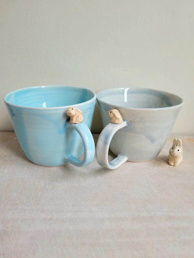 Two handmade pottery cups with blue and grey ceramic glaze with a bunny rabbit sitting on the handles