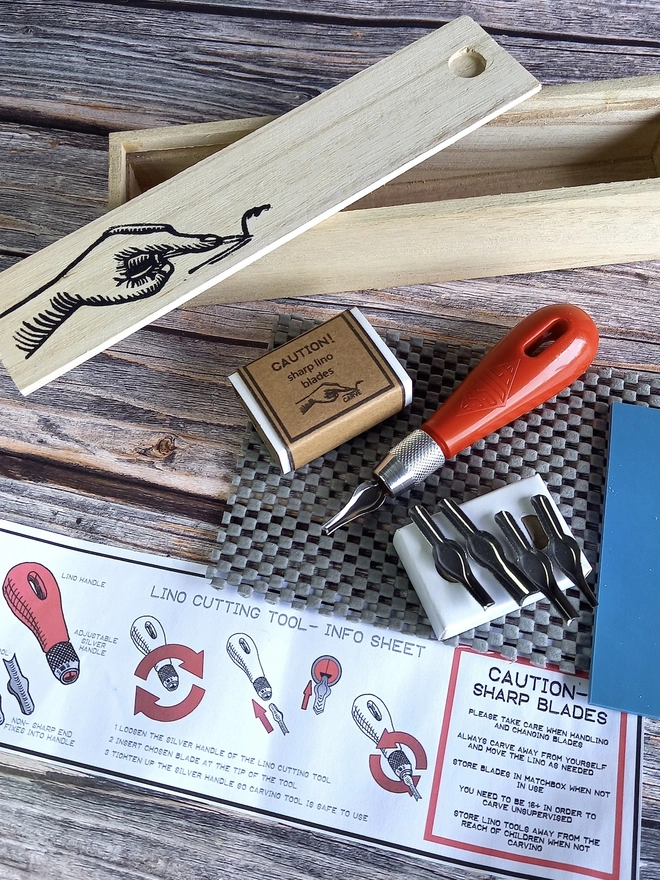Lino Carving Printing Tool Kit In Presentation Box, carving utensils and instruction sheet unpacked on a table next to wooden presentation box. 
