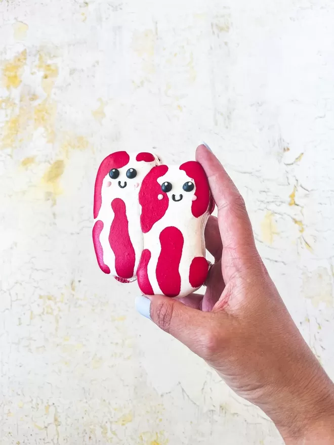 a hand holding two red and white rashers of bacon shaped  macarons with cute smiley faces