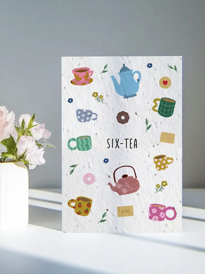 A decorative card titled “SIX-TEA” featuring colourful illustrations of teapots, cups, and small flowers, placed next to some pale pink flowers in soft natural light.