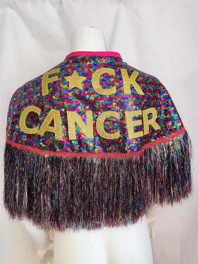 A cape sits on the back of a mannequin. The words 'FUCK CANCER' are across it but the 'U' has been replaced with a star to censor it. The sequins are rainbow as well as the tinsel, The neck tie is pink.