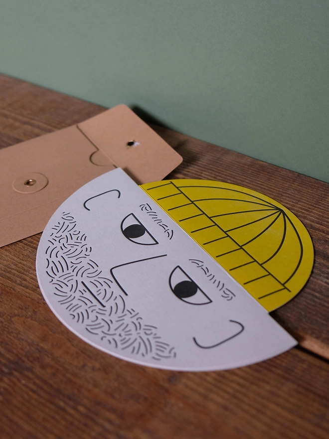 A round card with a male bearded face and yellow pop up hat. The card is hand letterpress printed in black and yellow ink on a light grey card. String and washer envelope in background. 