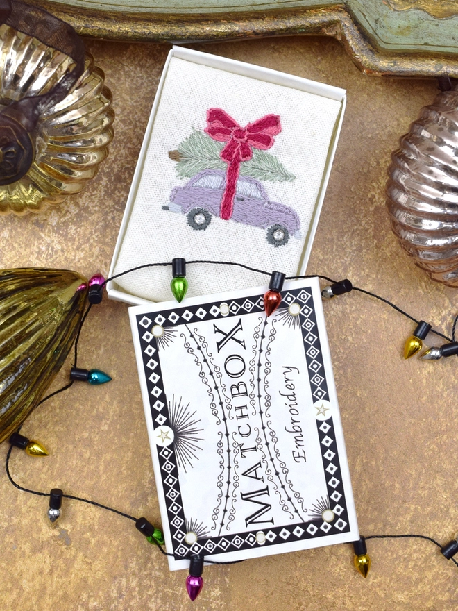 Christmas Tree Pick-Up by Car embroidery, inside a Matchbox displayed on a gold background with glass baubles.