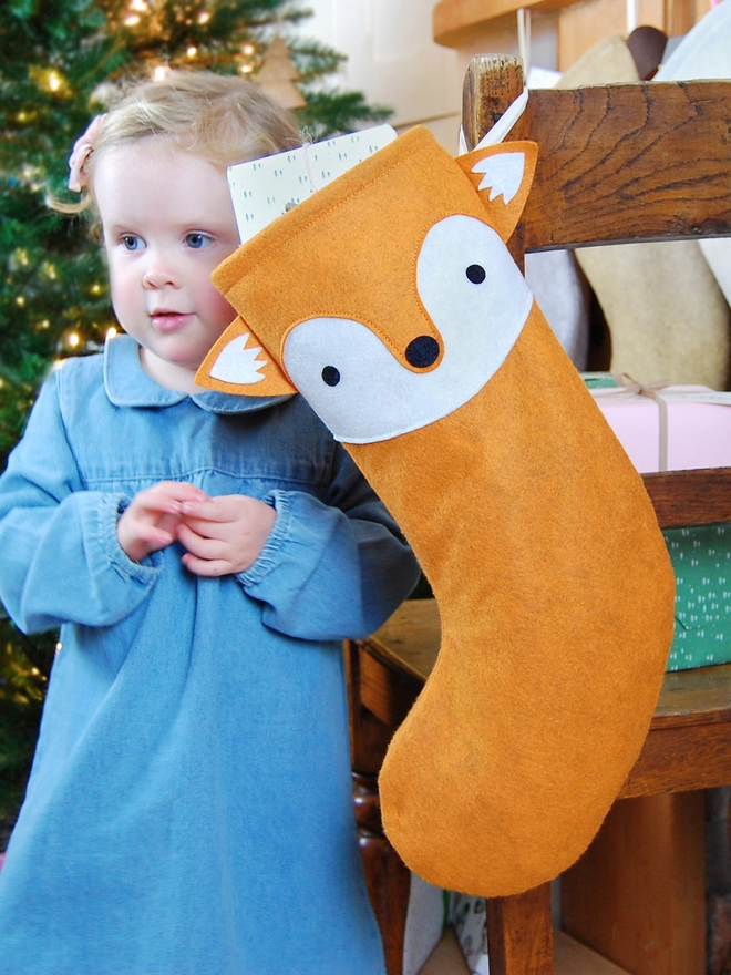 A handmade felt fox stocking hangs on a wooden chair, where a little girl sits, in front of a Christmas Tree.