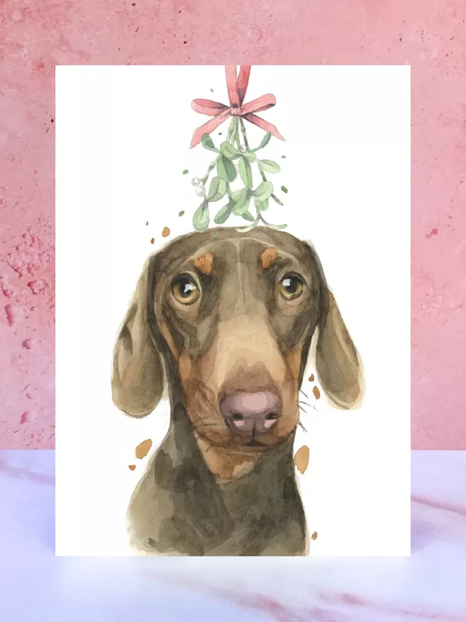 A Christmas card featuring a hand painted design of a Dachshund, stood upright on a marble surface.