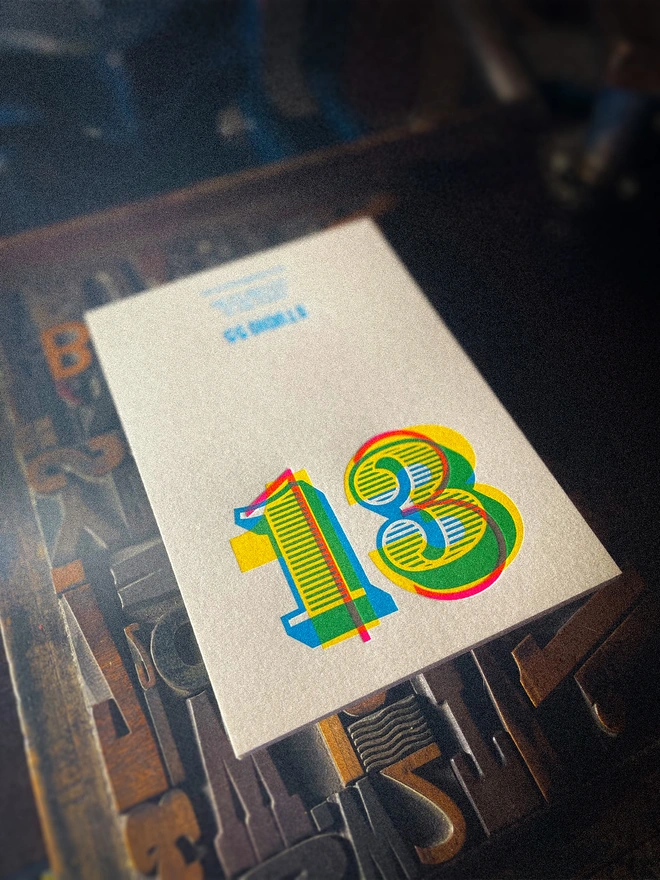 Handmade 13th birthday letterpress card shown open on vintage wooden printer's type, displaying the colourful overlapping number 13 design and Studio 55 Letterpress maker's mark, individually printed using multiple colour passes.
