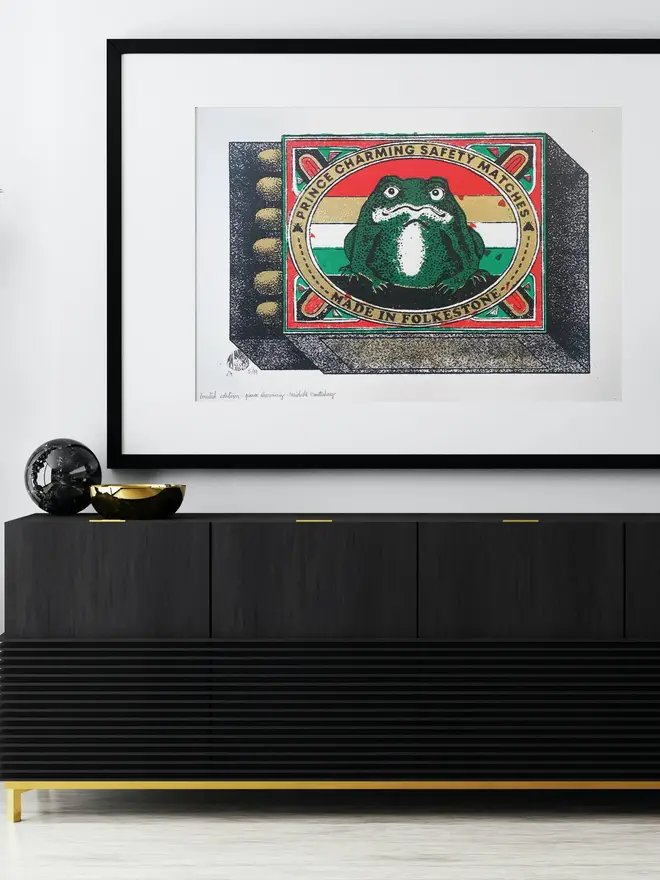  An image of a limited edition A2 screenprint. The print depicts a retro-style matchbox illustration with a central image of a green frog with a white belly, set against a background of horizontal red, white, and green stripes. The frog is framed by a gold-bordered oval. Text at the top of the oval reads "PRINCE CHARMING SAFETY MATCHES," and at the bottom, "MADE IN FOLKESTONE." The matchbox border is red with ornate details and a green trim. The match tips on the left are a textured gold. Below the main image, the print is signed and numbered 5/11. The entire image has a grainy texture, characteristic of a vintage screenprint.