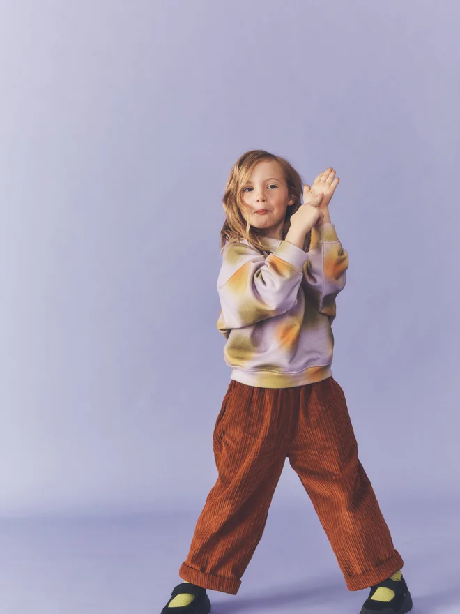 Overhead shot of a child laughing and holding beads, wearing the Studio Koter Luka jersey top in Hazy Lilac against a lilac backdrop.