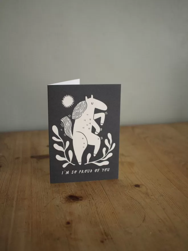 Black and white greeting card with illustration and the words I'm so proud of you written on it stood up on wooden surface 