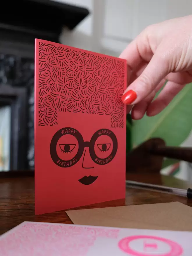 Coral card with a ladies face wearing glasses saying ' Happy Birthday' and big square hair. 