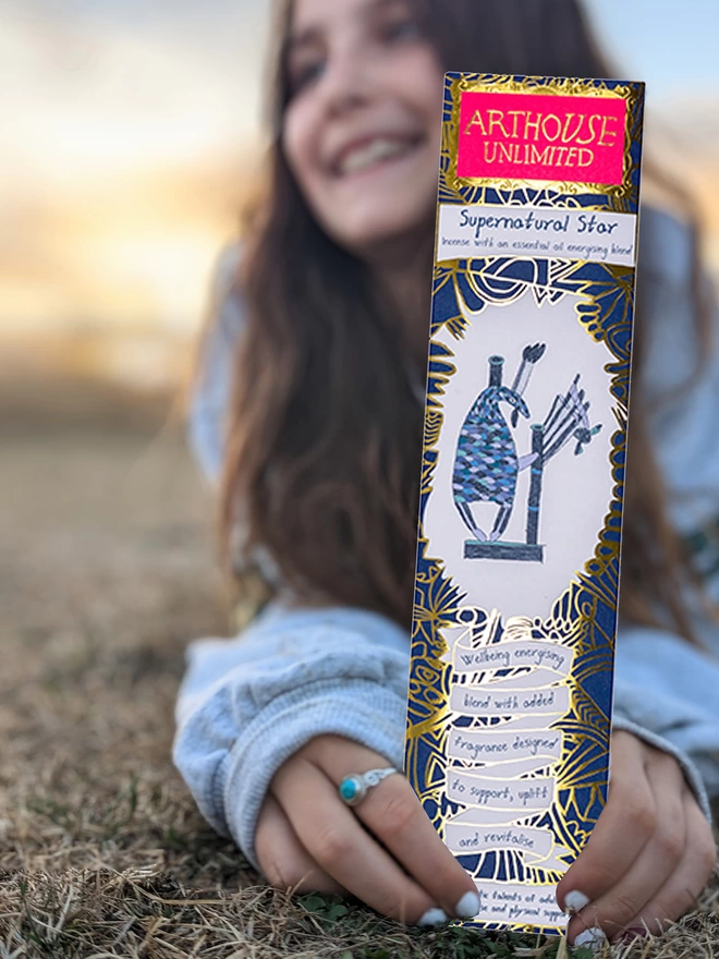 happy girl in a field holding supernatural star well being charity incense sticks with blue illustrations