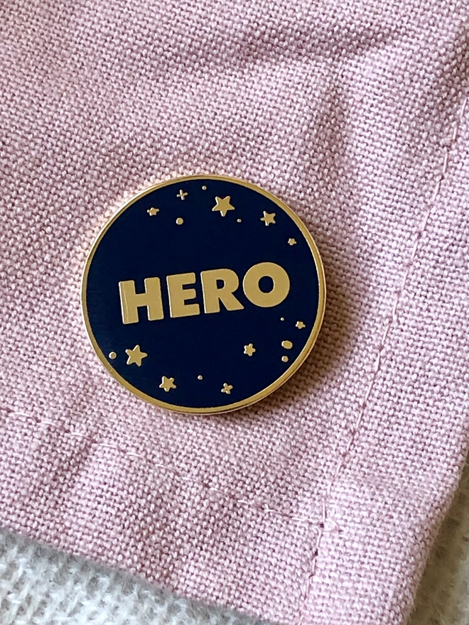 A navy blue and gold enamel pin badge with a starry design and the word "Hero" is pinned to a pink blanket.