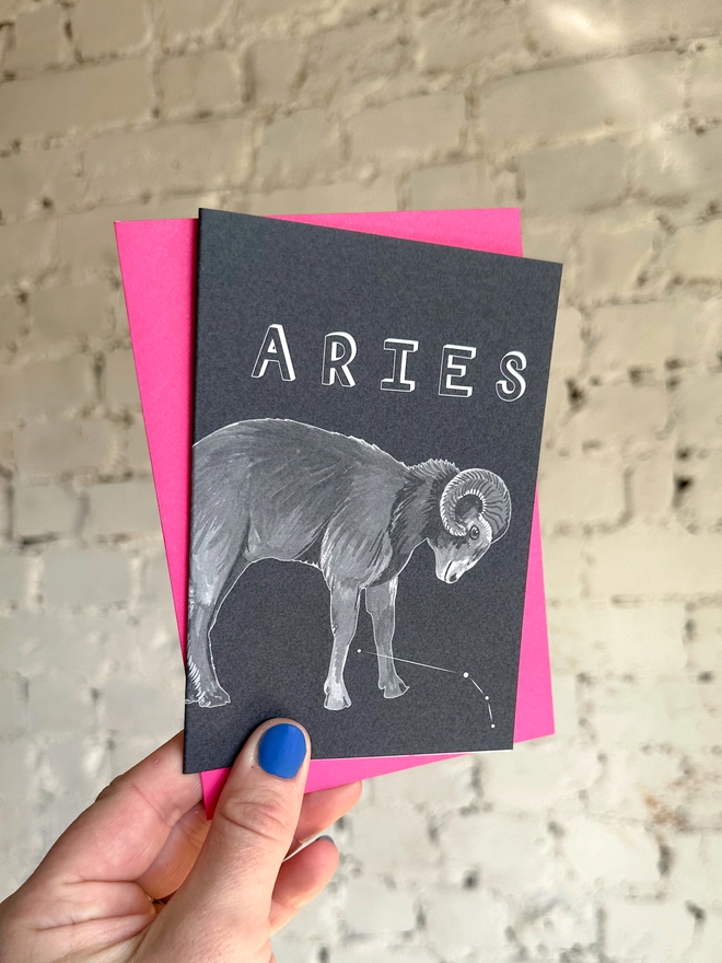 a greetings card featuring a greyscale illustration of a zodiac style ram with the text “ARIES” with a neon pink envelope