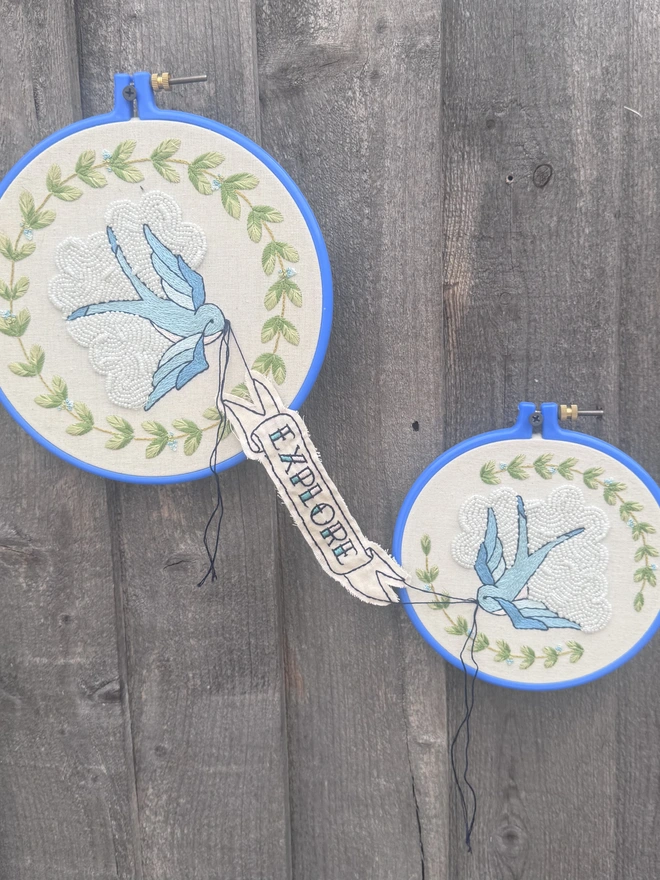 Two blue embroidery hoops containing blue swallows holding a banner between them saying explore  
