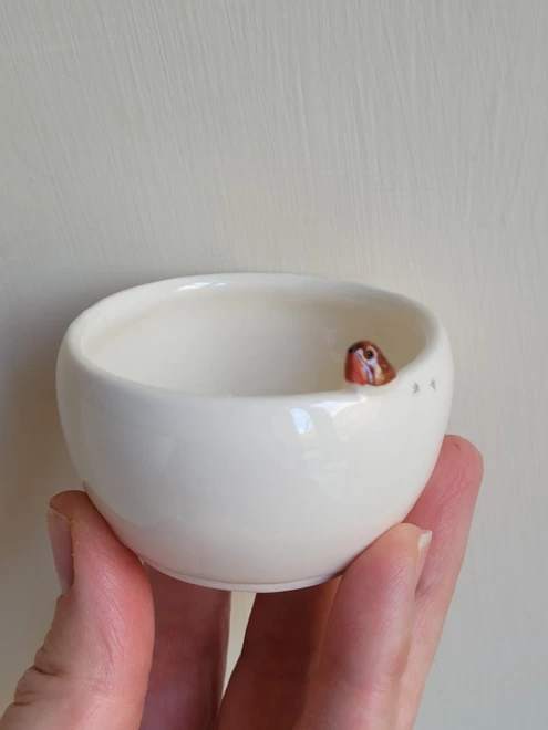 hand holding aloft a small ceramic white pot with a little modelled robin on the edge and tiny foorprints to the side