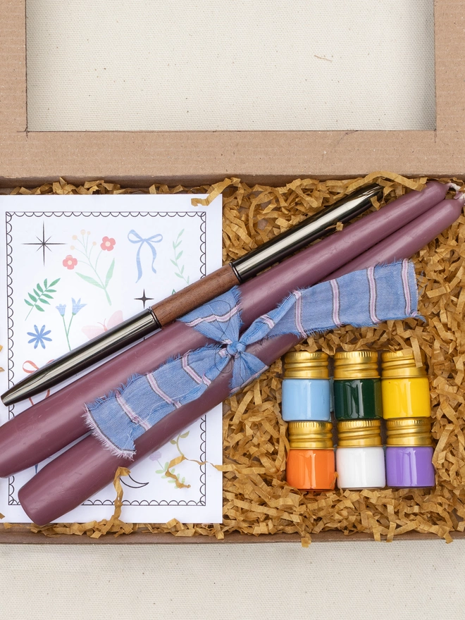 Open candle painting kit showing two mauve taper candles, six colourful candle paints, a double-ended paintbrush and an illustrated instruction booklet nestled in a kraft gift box with shredded paper filling.