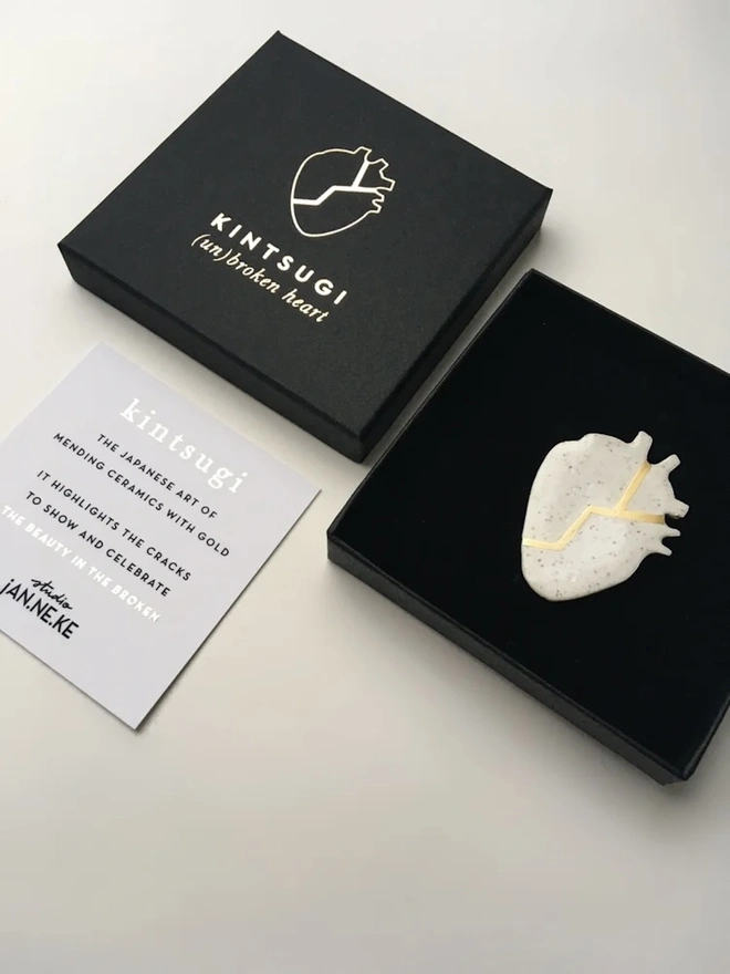 (Un)Broken Heart Pin, a white and gold heart pin sitting on a white surface next to its black box and card. 