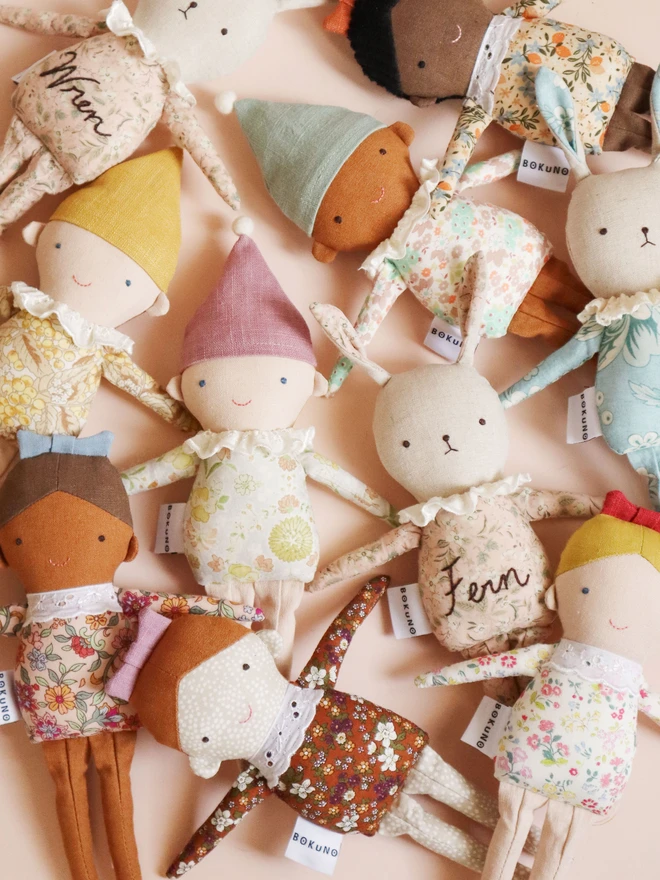 Three handmade Spring Elf Dolls are sitting side by side against a soft pastel background. Each doll has a floral print body, pastel-coloured hat with a pompom, and a ruffled white collar. They feature different skin tones and gentle smiles, radiating charm and warmth.