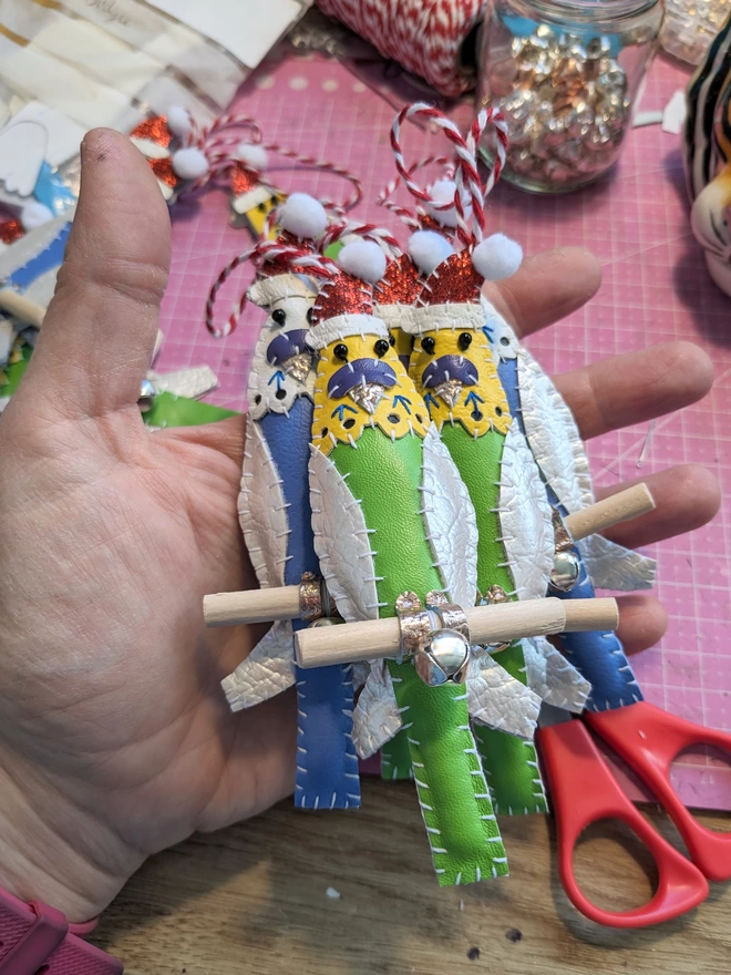 A handful of hand stitched faux leather budgie Christmas decorations wearing Santa hats. One bird is blue, one green.