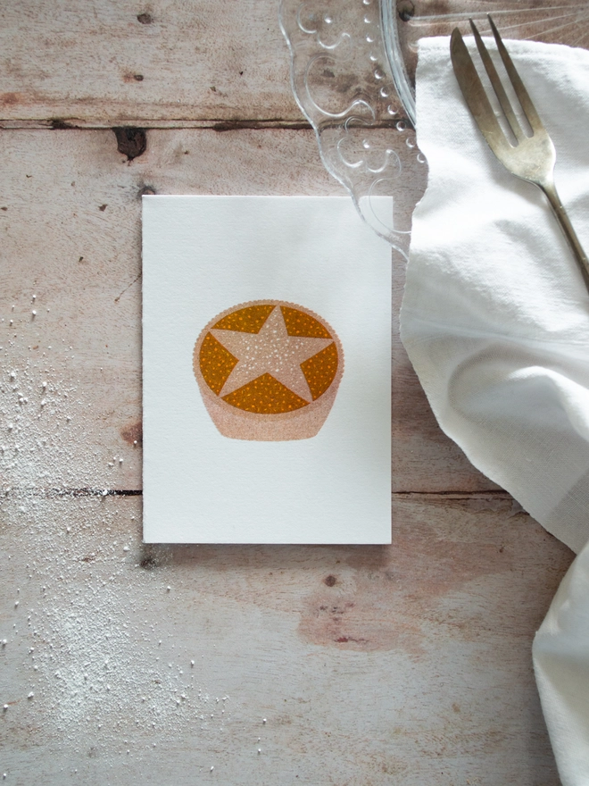 A small festive Christmas crd printed with an illustrated Mince pie lies on a wooden Christmas table beside and empty plate, napkin and desert fork.